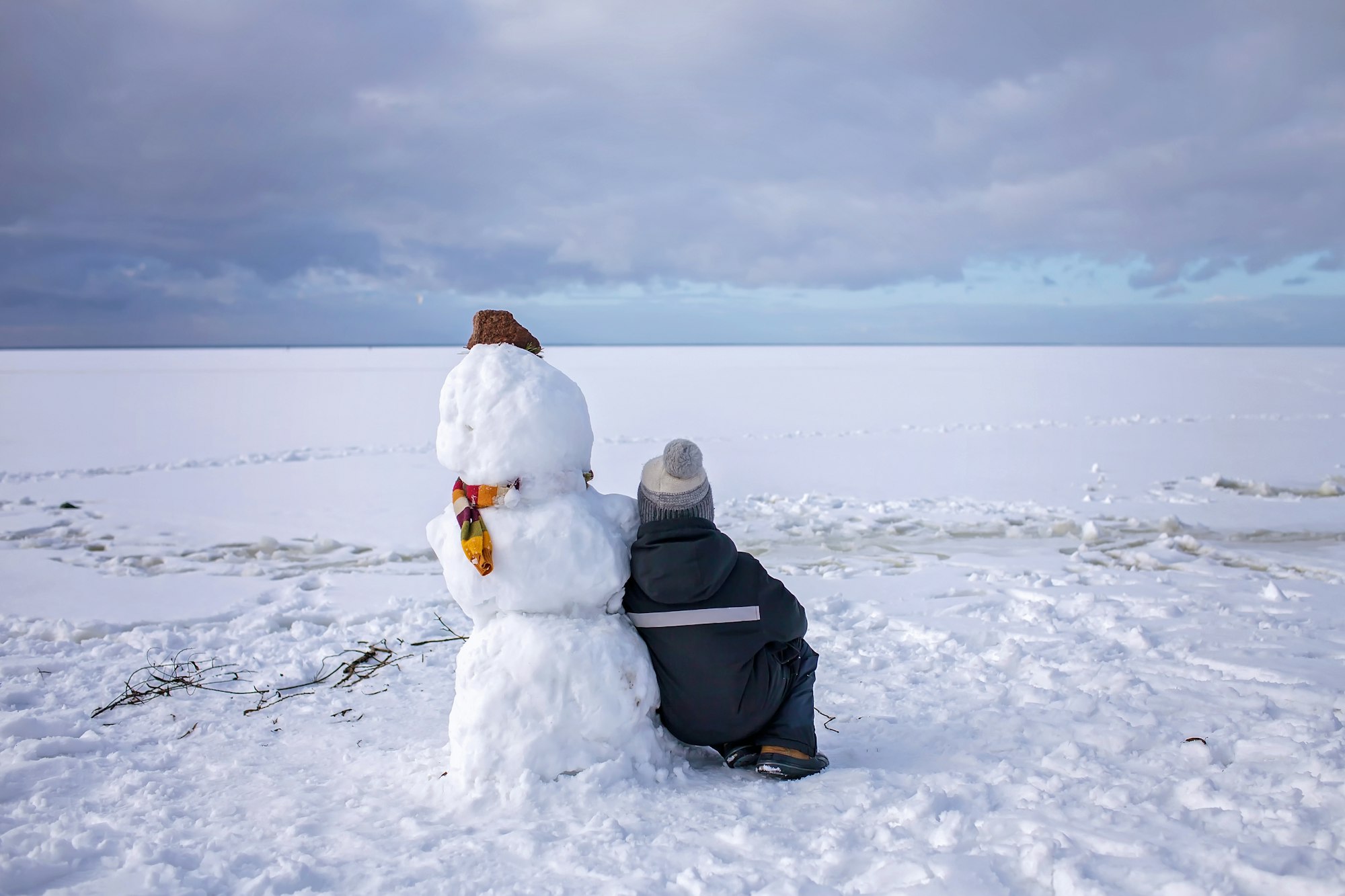 Boy sits alone with snowman and looks forward. Loneliness and friend made of snow. Winter mood